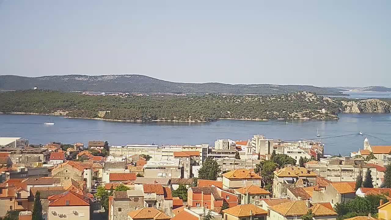 A coastal town with red-tiled roofs sits beside a calm body of water, with a lush green island in the distance under a clear sky.