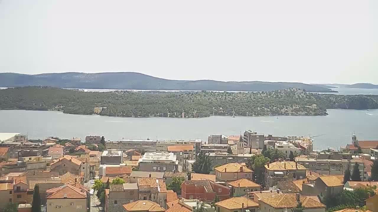A town with orange-tiled roofs overlooks a calm body of water and a verdant island under a bright, clear sky.
