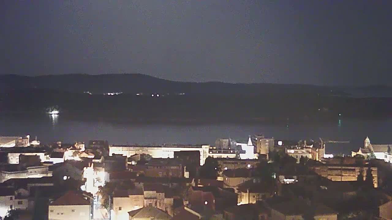 A nighttime view of a town situated on a body of water, with dimly lit buildings and a dark, mountainous backdrop under a clear sky.