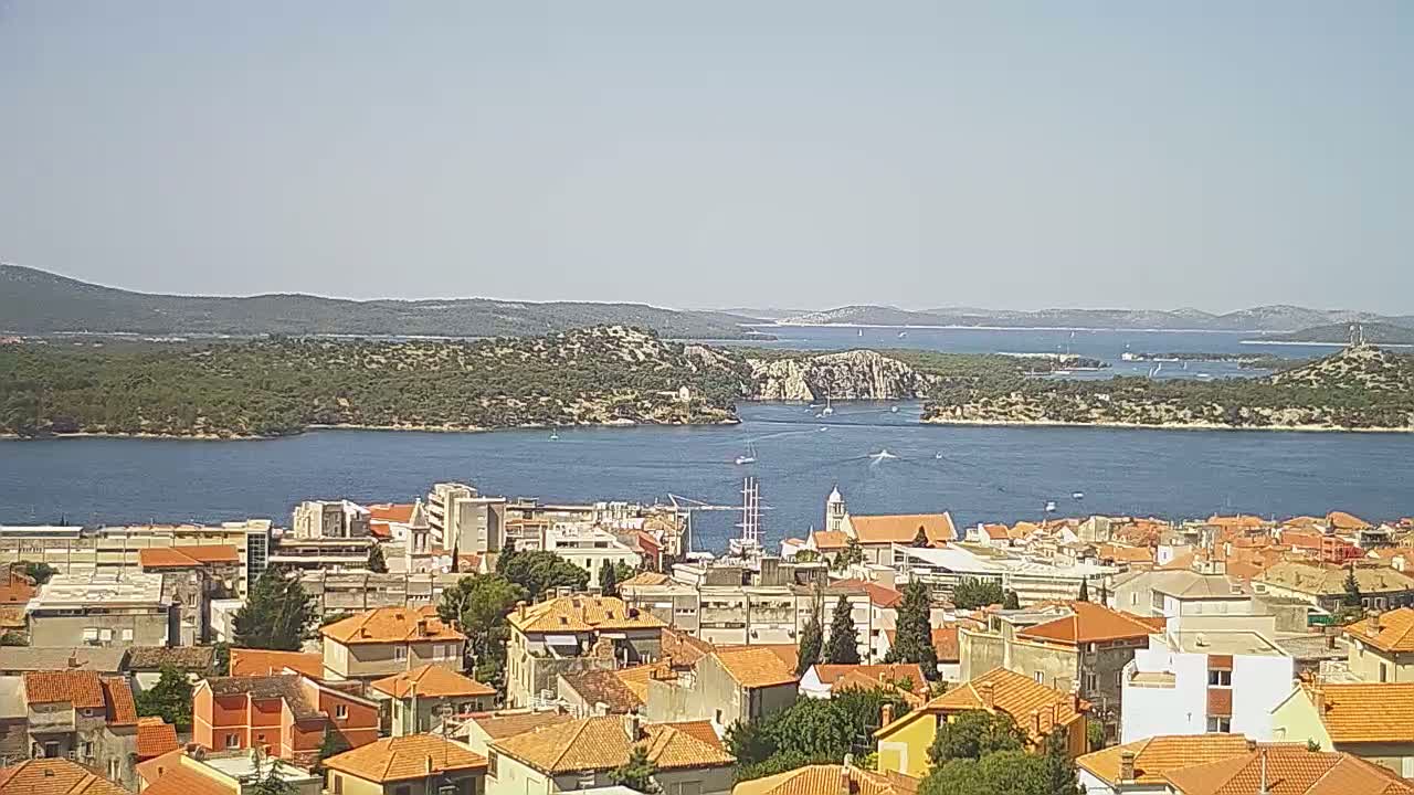 A coastal town with orange-roofed buildings overlooks a calm blue bay surrounded by green islands on a clear, sunny day.