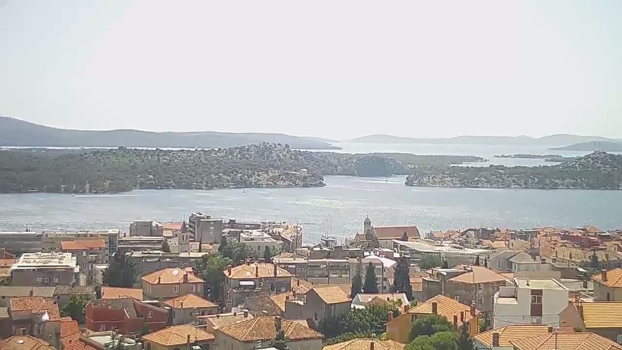 A coastal town with red-tiled roofs overlooks a calm, sunlit body of water and several islands on a clear day.