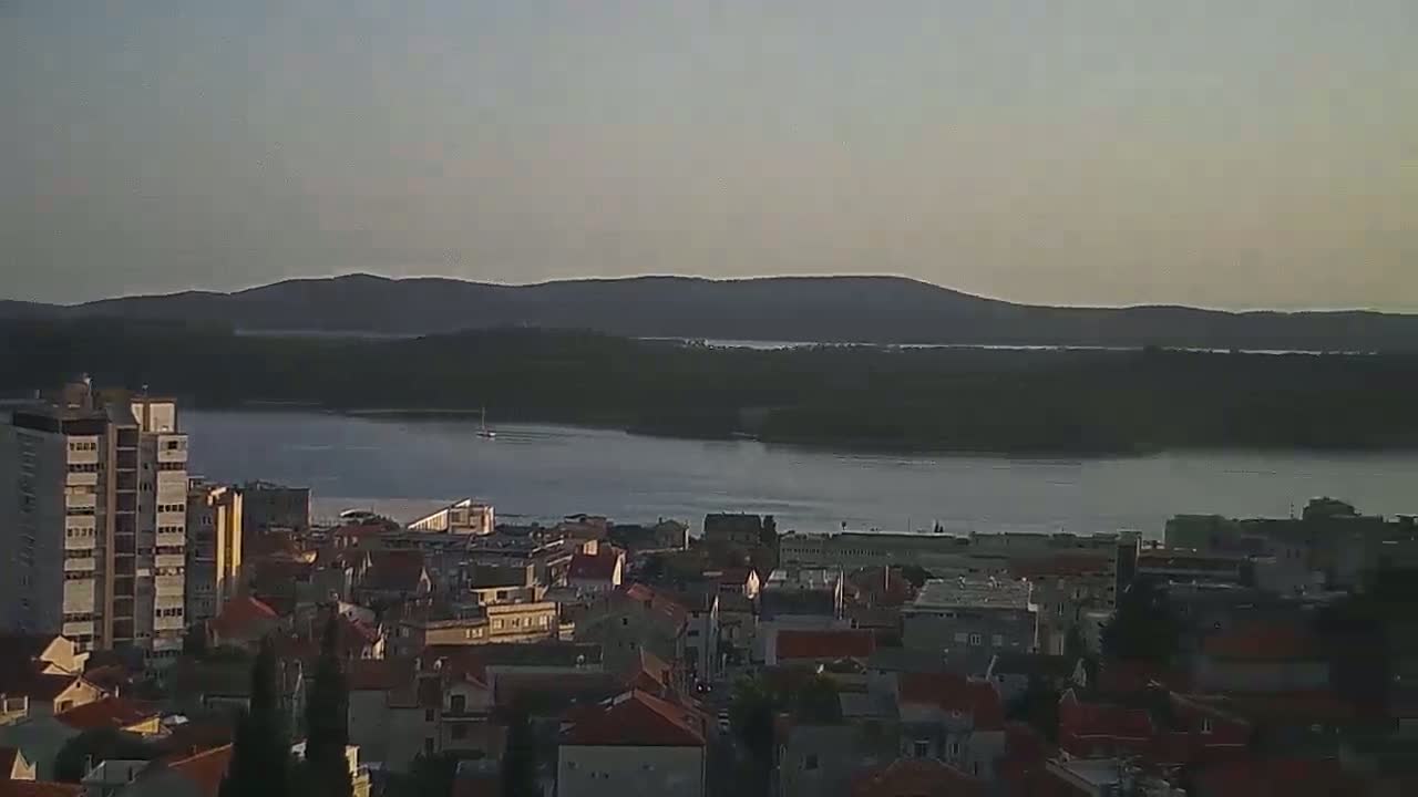 A coastal city with low-rise buildings and a few high-rises overlooks a calm body of water, beyond which lie dark, hilly lands under a clear, dusky sky.