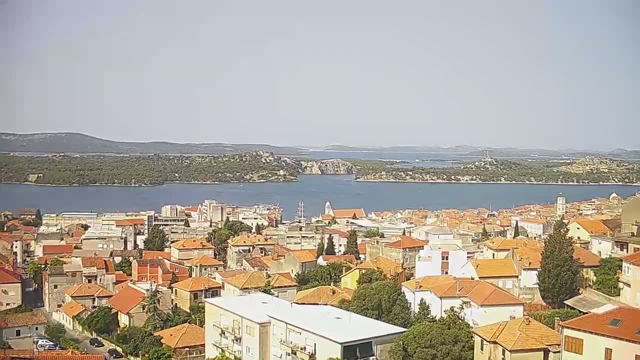 A coastal town with red-tiled roofs is situated on a hill overlooking a calm body of water under a clear sky.