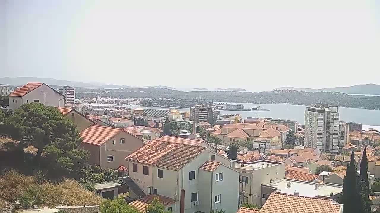 A hazy view of a coastal town with red-tiled rooftops, situated on a hillside overlooking a calm body of water and distant islands.