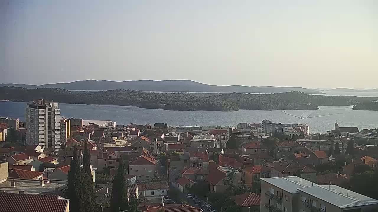 A coastal town with red-roofed buildings overlooks a calm body of water and distant islands under a clear, bright sky.