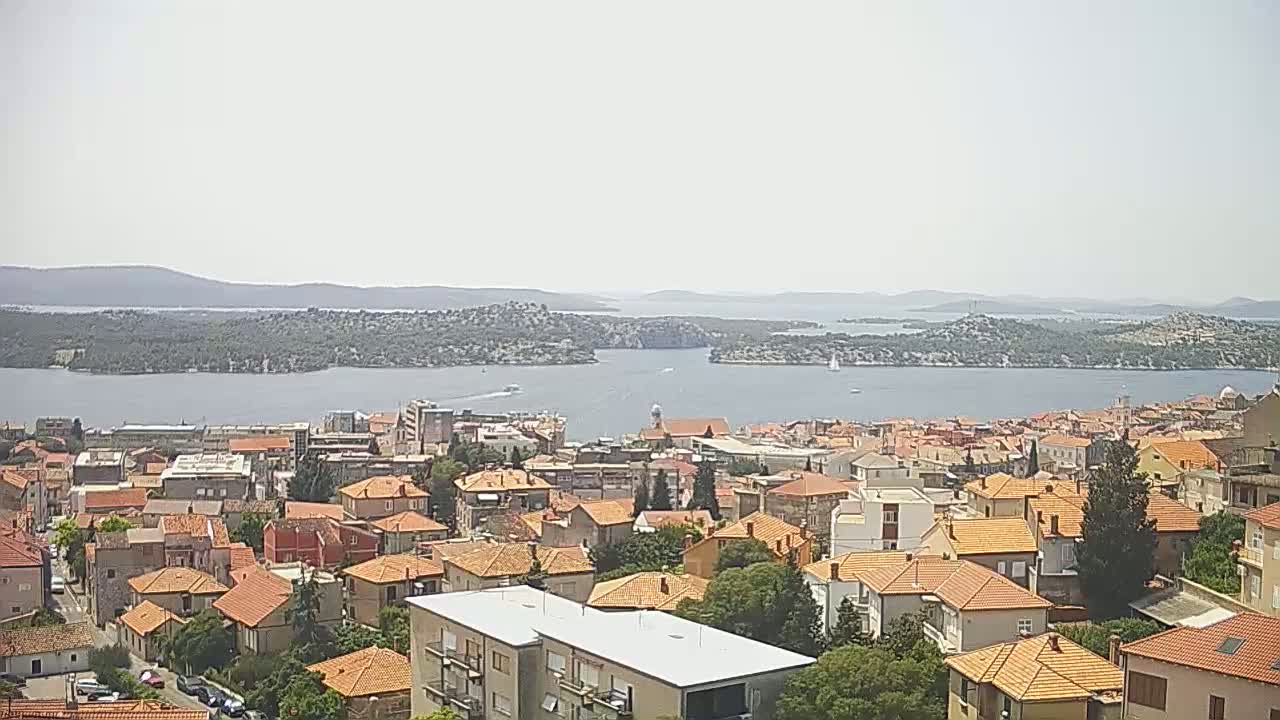 A coastal town with red-tiled roofs overlooks a calm bay and distant islands under a hazy sky.