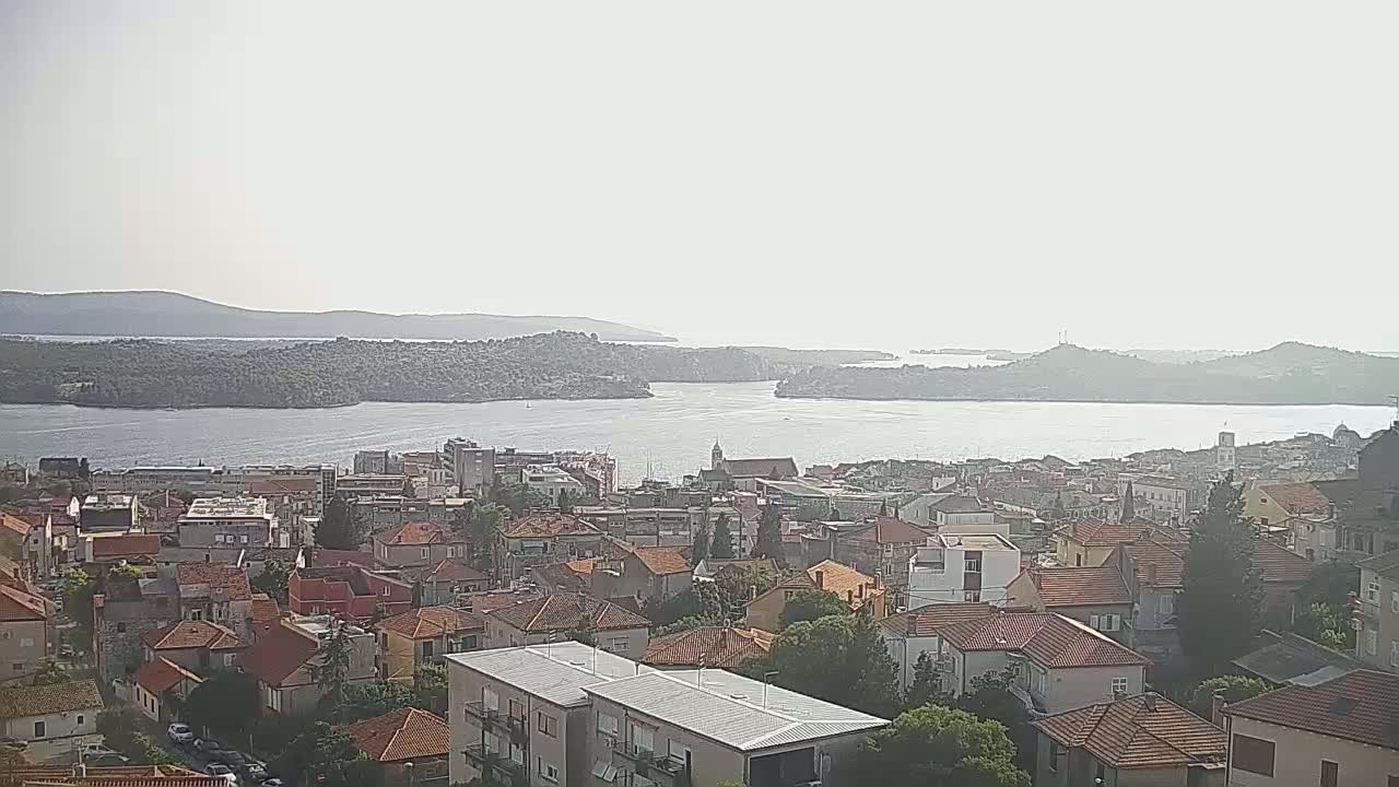 A hazy, sunny day reveals a coastal town with red-tiled roofs and a calm body of water separating it from distant, tree-covered hills.