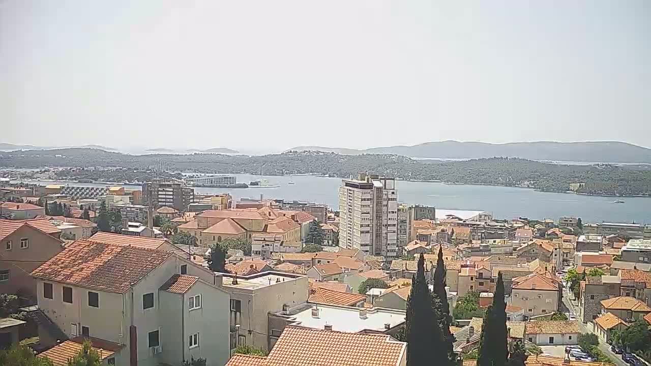 A sunny view of a coastal town with red-tiled roofs, a multi-story building, and a body of water with islands in the distance.