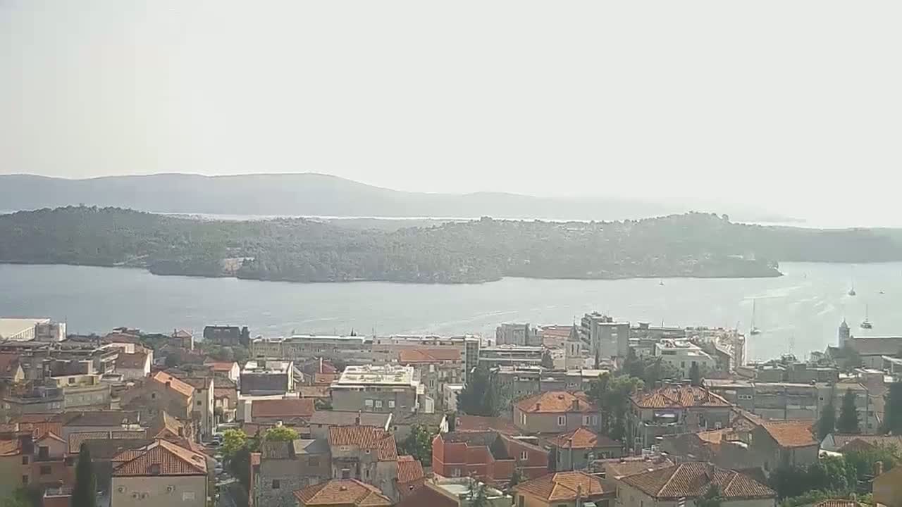 A hazy, sunny day overlooks a coastal town with red-tiled roofs, a body of water, and distant islands.