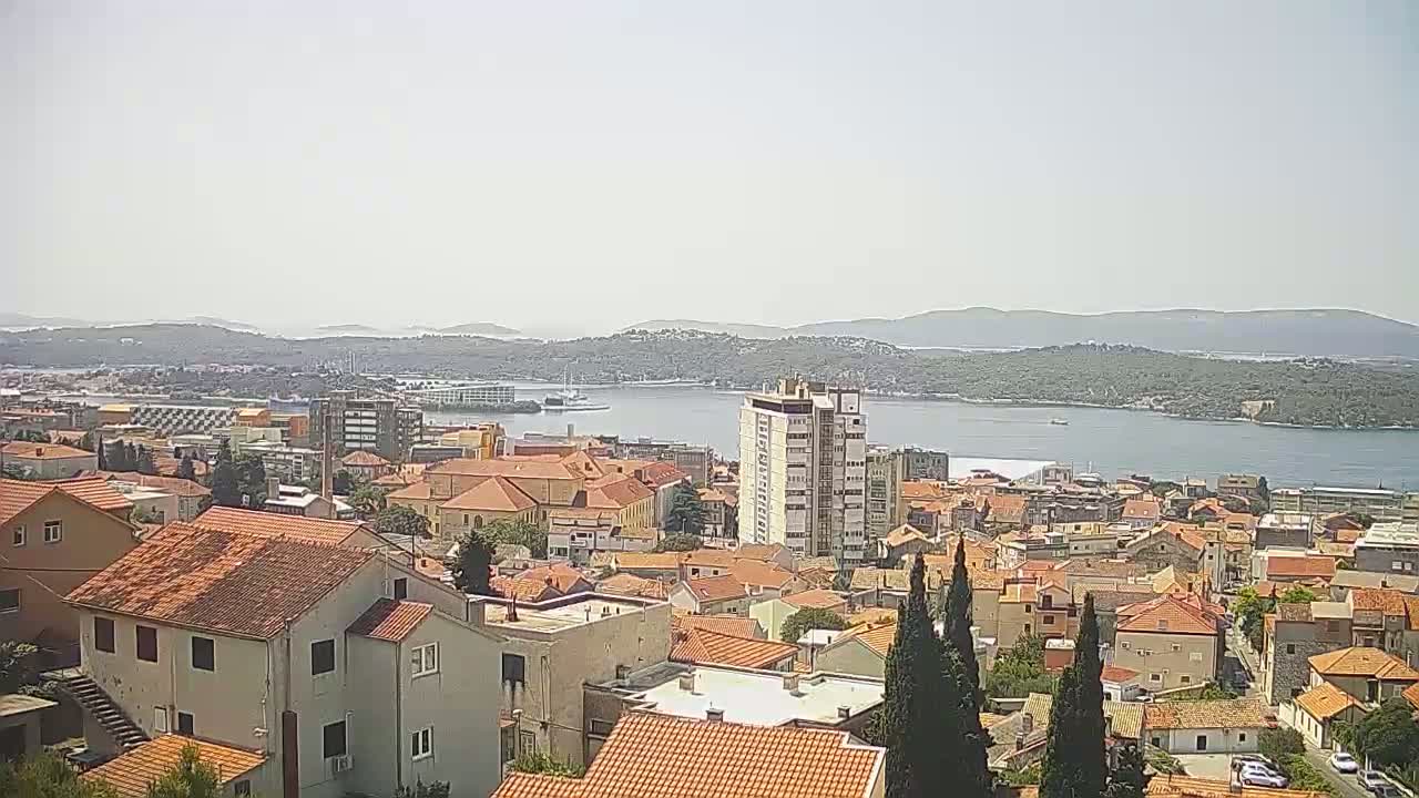 A sunny view of a coastal town with red-tiled roofs, a tall building, and a body of water with islands in the distance.