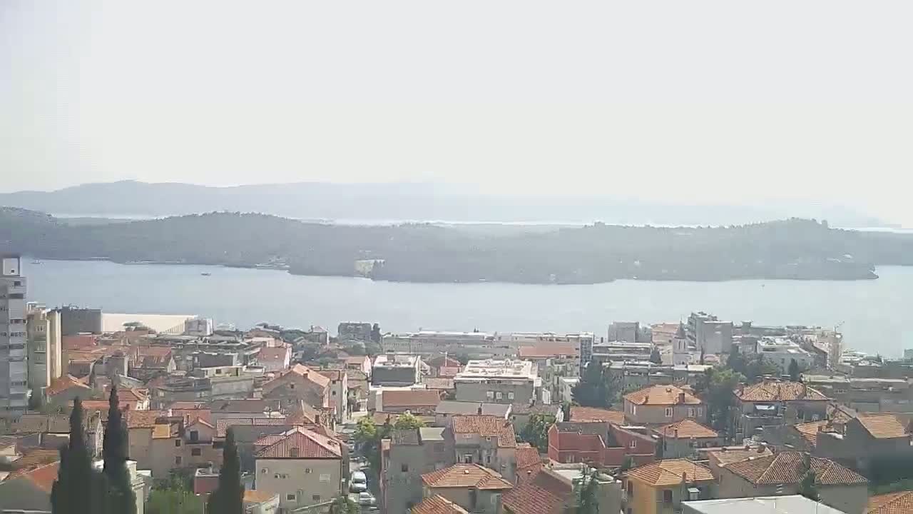 A hazy view shows a coastal town with red-tiled roofs overlooking a calm body of water and distant islands.