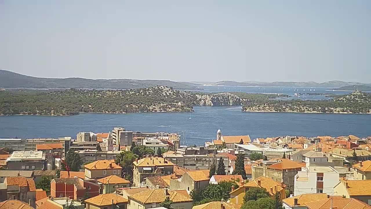 A coastal town with orange-tiled roofs overlooks a calm blue body of water,  surrounded by green hills and distant islands under a clear sky.