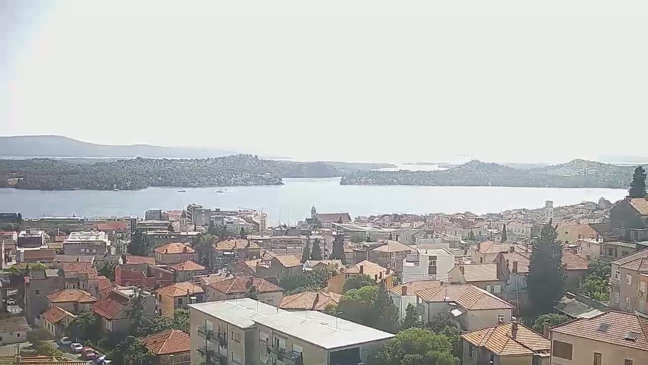 A hazy, sunny day overlooks a coastal town with red-tiled roofs, nestled beside a calm body of water and surrounded by distant islands.