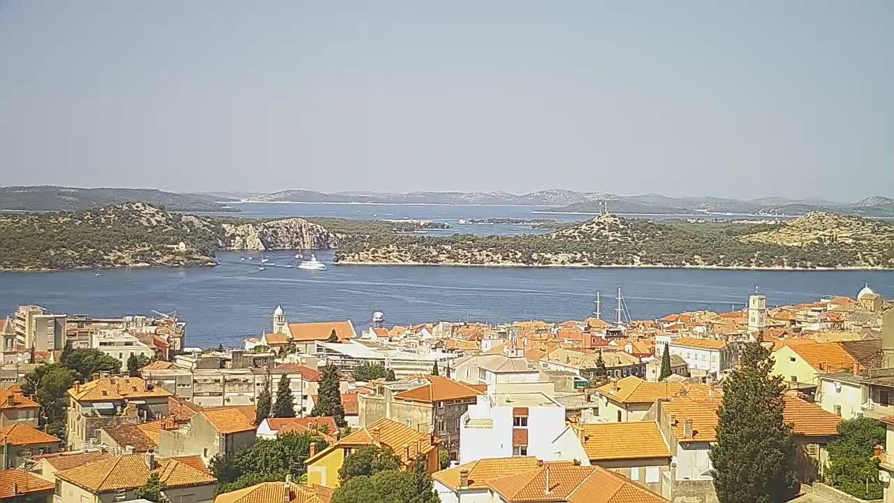 A coastal town with orange-tiled roofs overlooks a calm body of water dotted with islands under a clear, sunny sky.