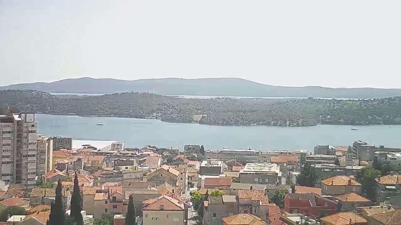 A coastal town with red-tiled roofs overlooks a calm bay and distant hills under a hazy sky.