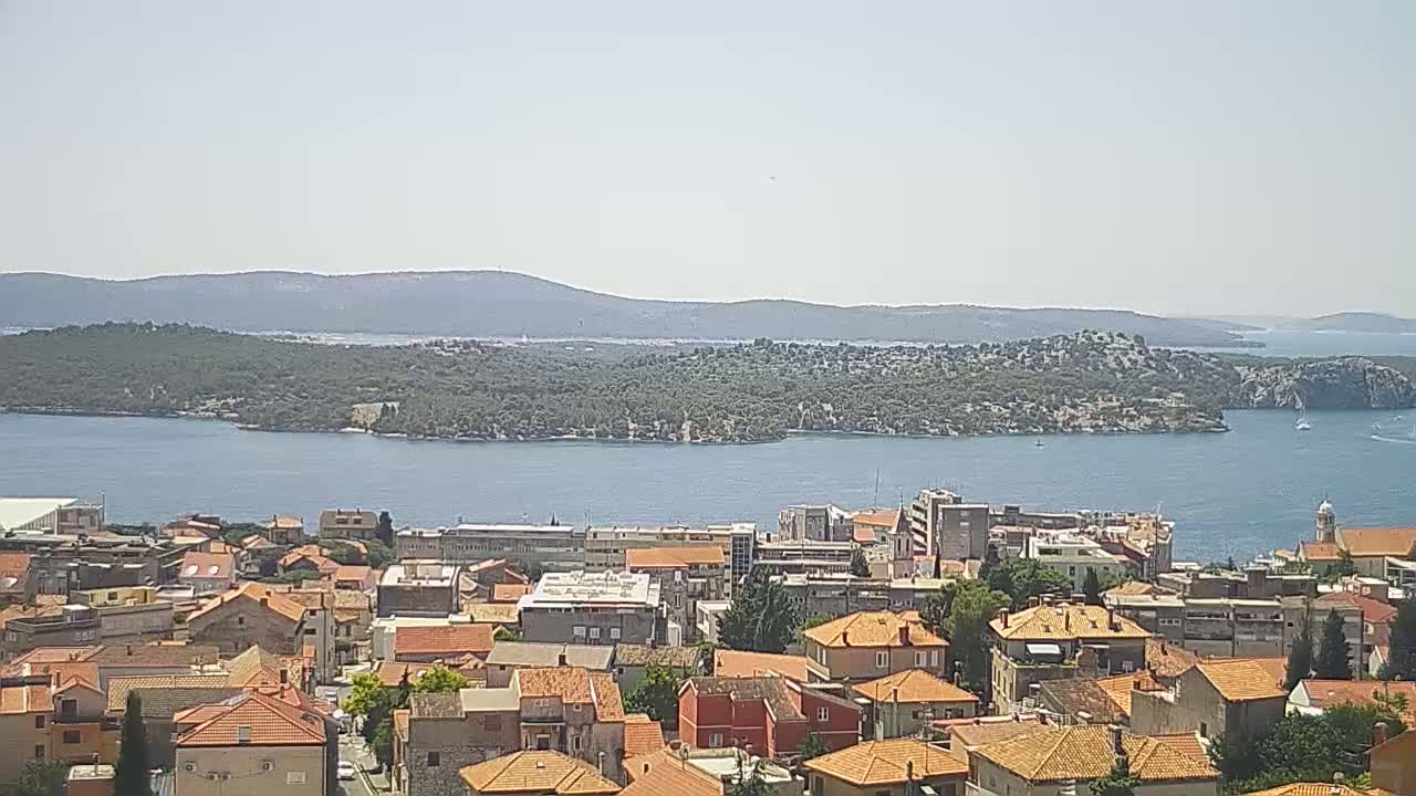 A coastal town with red-tiled roofs overlooks a calm bay and a verdant island under a clear, sunny sky.