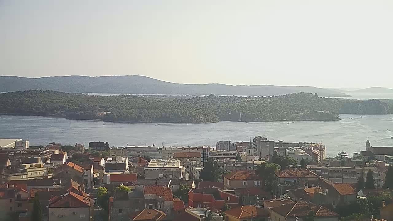 A hazy, sunny day reveals a city of red-roofed buildings overlooking a calm body of water with an island in the distance and mountains beyond.