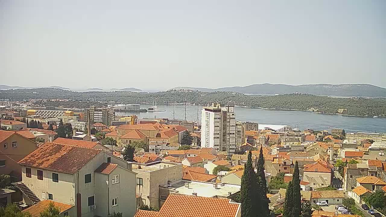A coastal town with red-tiled roofs, a multi-story building, and a body of water with islands in the background on a clear, sunny day.