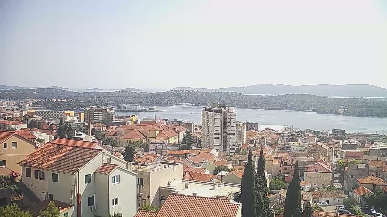 A coastal city with red-tiled roofs and a high-rise building is visible, under a clear, sunny sky, overlooking a calm body of water and distant hills.