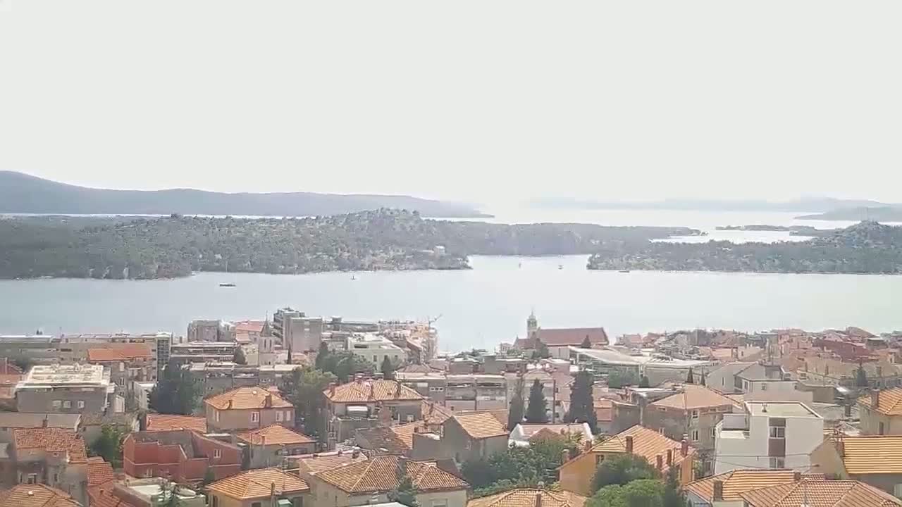A coastal town with red-tiled roofs overlooks a calm body of water and distant islands under a hazy sky.