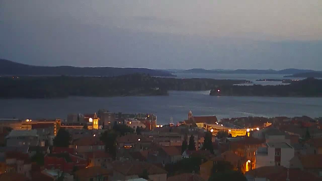 A coastal town at twilight, illuminated by scattered lights, overlooks a calm body of water dotted with islands under a dusky sky.