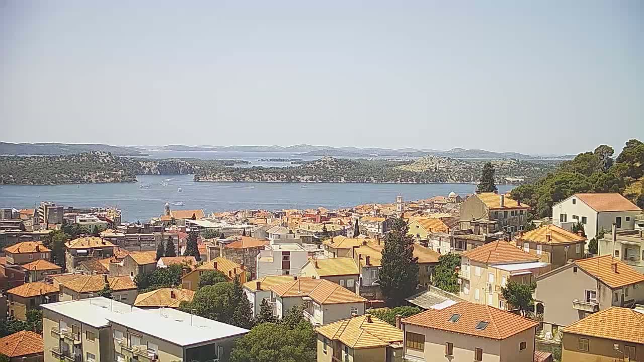 A coastal town with red-tiled roofs stretches toward a calm body of water under a clear, sunny sky, with islands and hills visible in the distance.