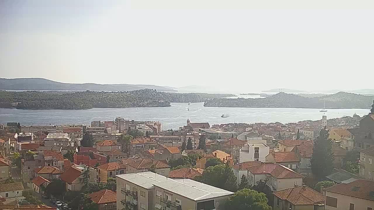 A hazy, sunny day reveals a coastal town with red-tiled roofs, nestled beside a calm body of water dotted with islands and boats, in the background.