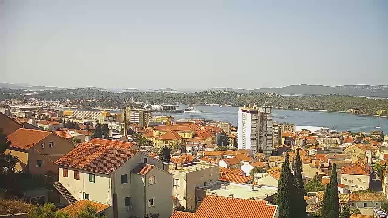 An elevated view of a coastal town with red-tiled roofs, a body of water, and distant hills under sunny skies.