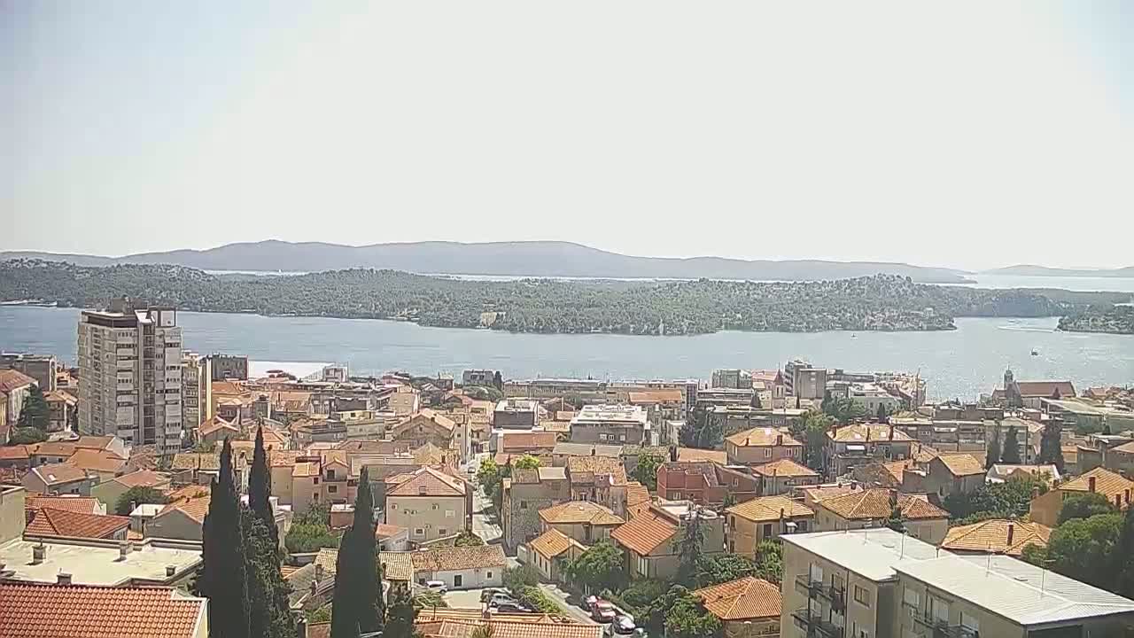 A coastal town with red-tiled roofs stretches to a calm body of water under a bright, sunny sky, with a mountainous island visible in the distance.