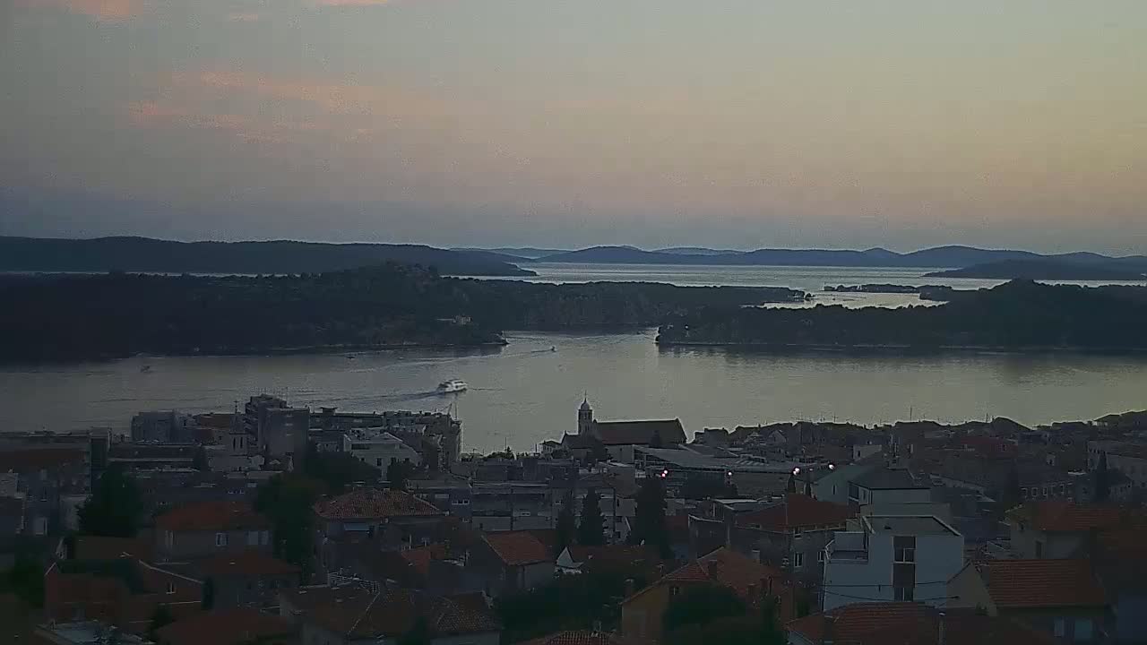 A coastal town with red-tiled roofs sits below a calm body of water at dusk, with dark silhouettes of islands and hills in the background under a pale sky.