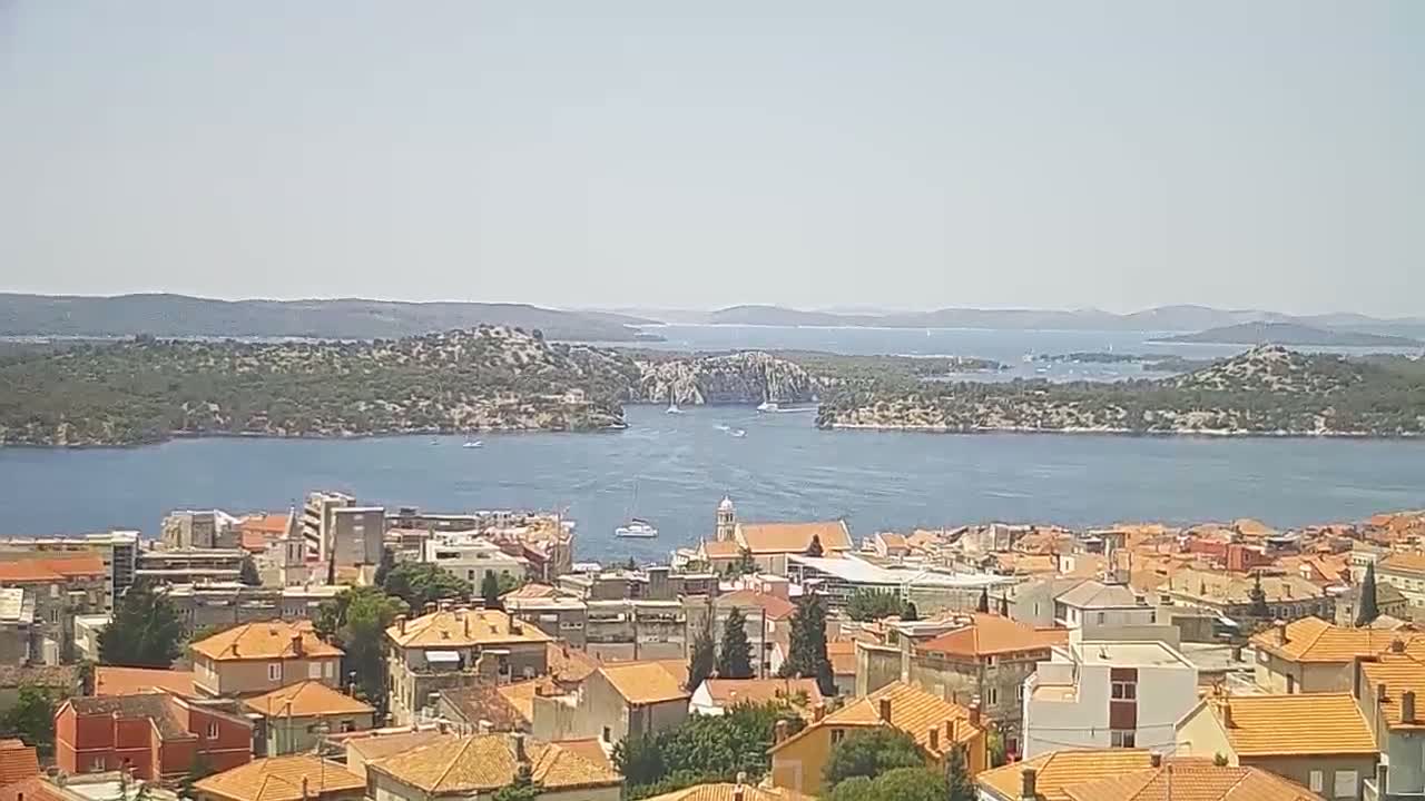 A coastal town with orange-tiled rooftops overlooks a calm blue bay dotted with islands under a clear sky.