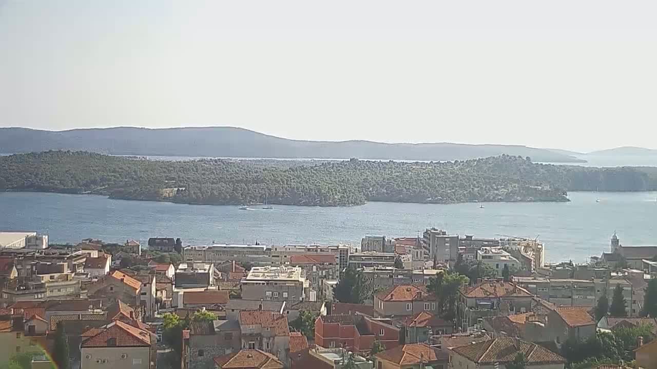 A coastal town with red-tiled roofs overlooks a calm body of water, with an island and distant mountains visible under a clear sky.