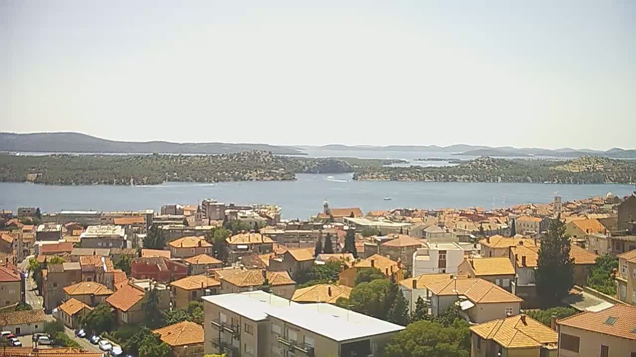 A coastal town with terracotta-roofed houses overlooks a calm bay and distant islands under a bright, clear sky.