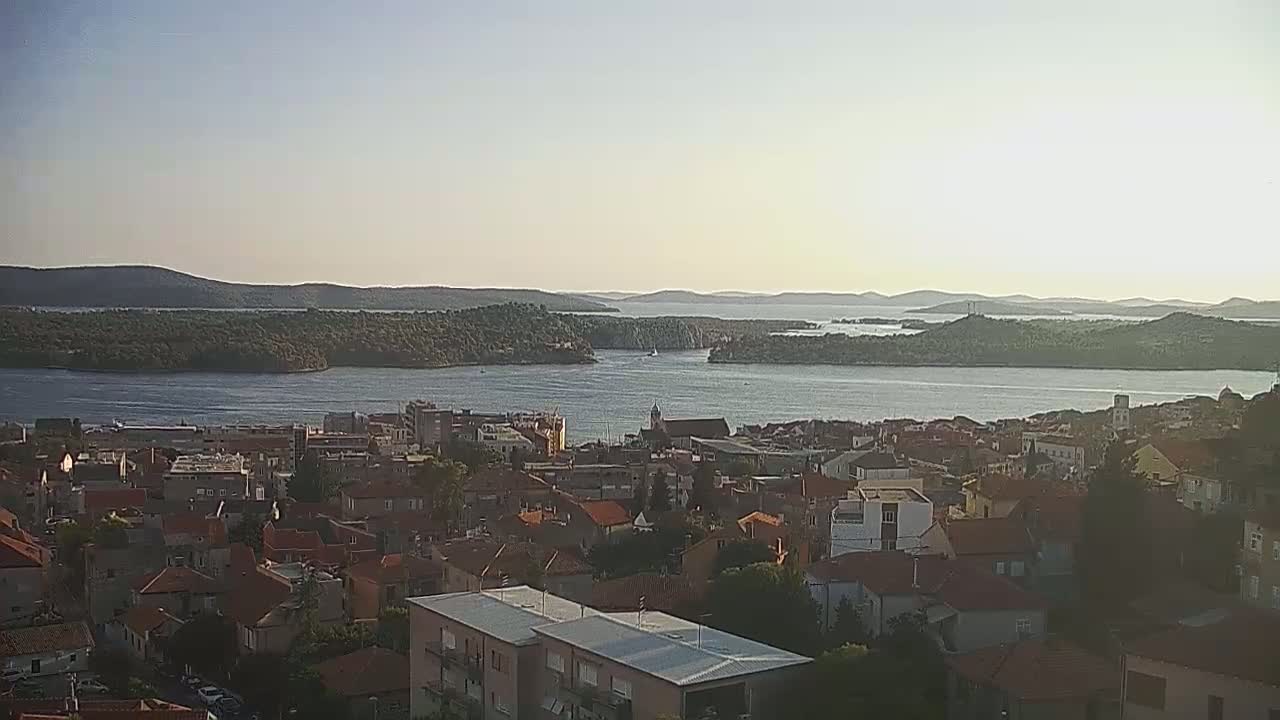 A coastal town with red-tiled roofs overlooks a calm bay and distant islands under a clear, bright sky.