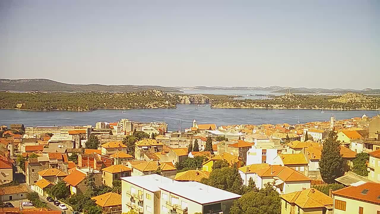 A coastal town with predominantly orange-roofed buildings sits on a waterfront, under a clear, sunny sky, overlooking a calm body of water and distant islands.