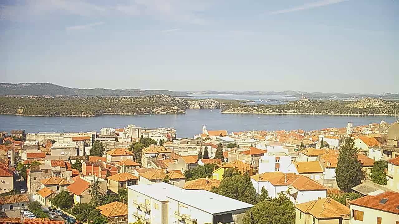 A coastal town with orange-tiled roofs sits on a hillside overlooking a calm bay and distant islands under a clear sky.