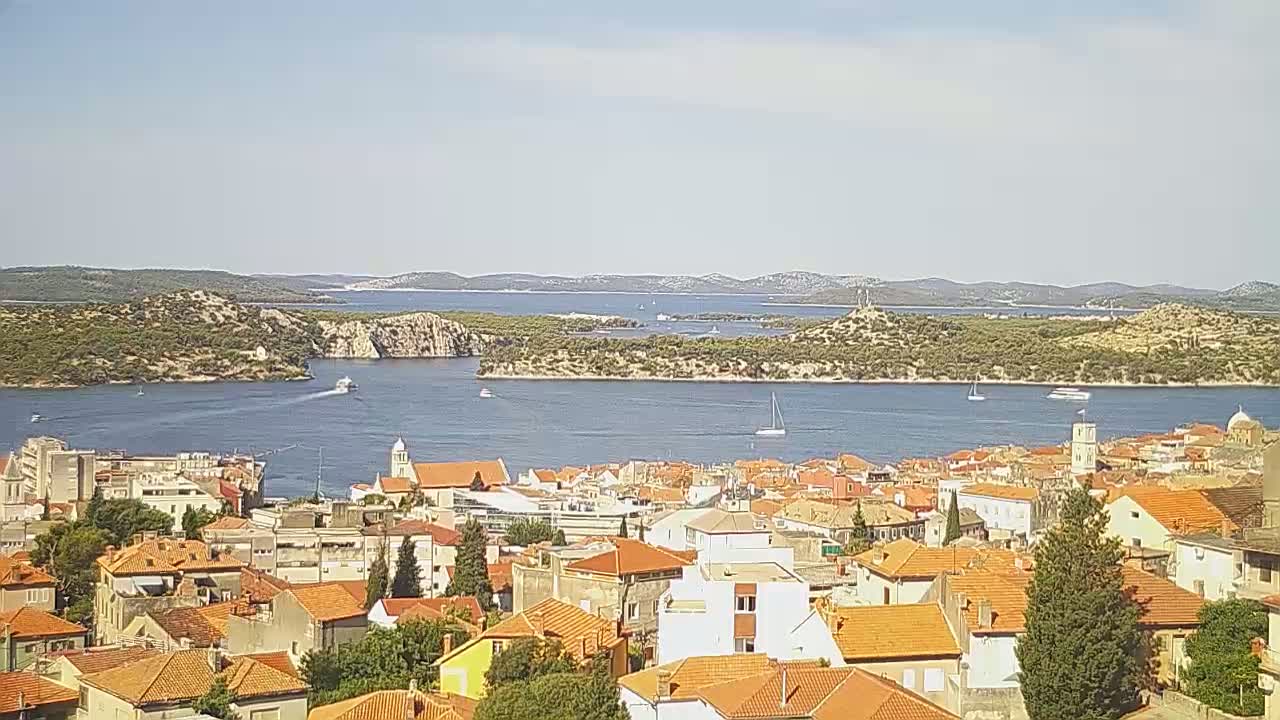 A coastal town with orange-tiled roofs overlooks a calm blue bay dotted with boats, under a clear, sunny sky.