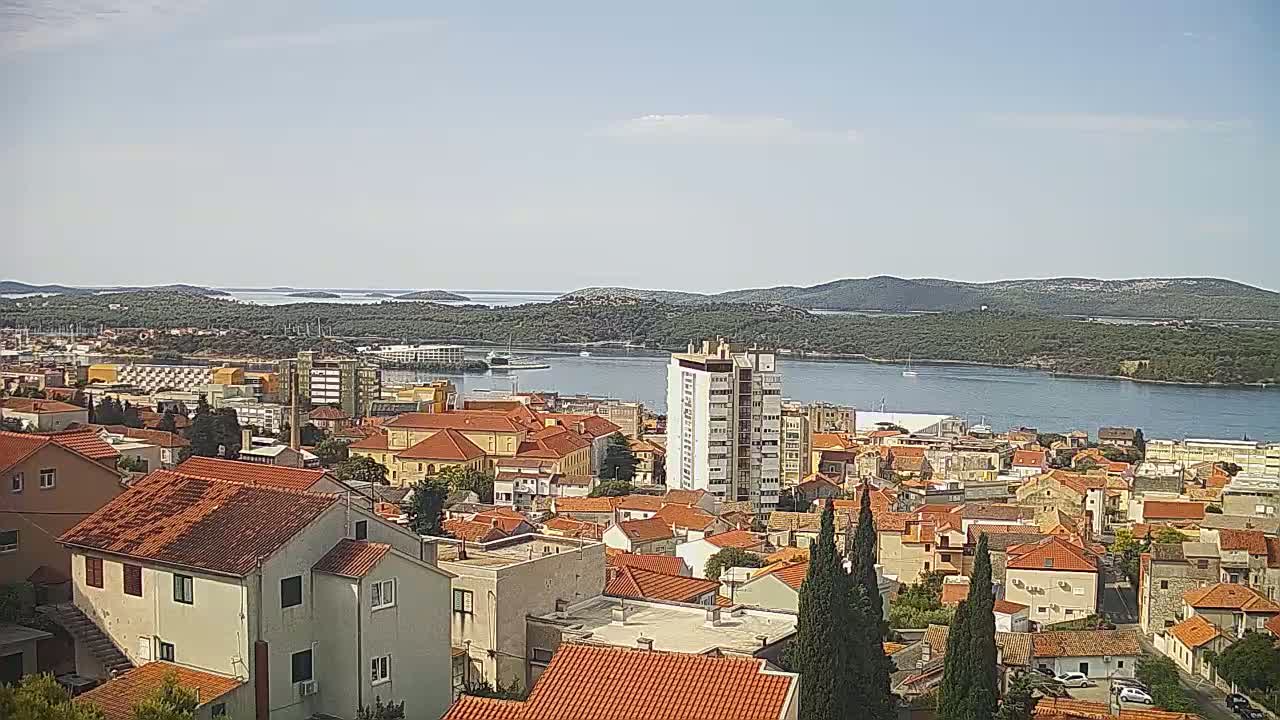 A coastal town with red-tiled roofs, a multi-story building, and a calm body of water surrounded by green hills under a mostly clear sky.