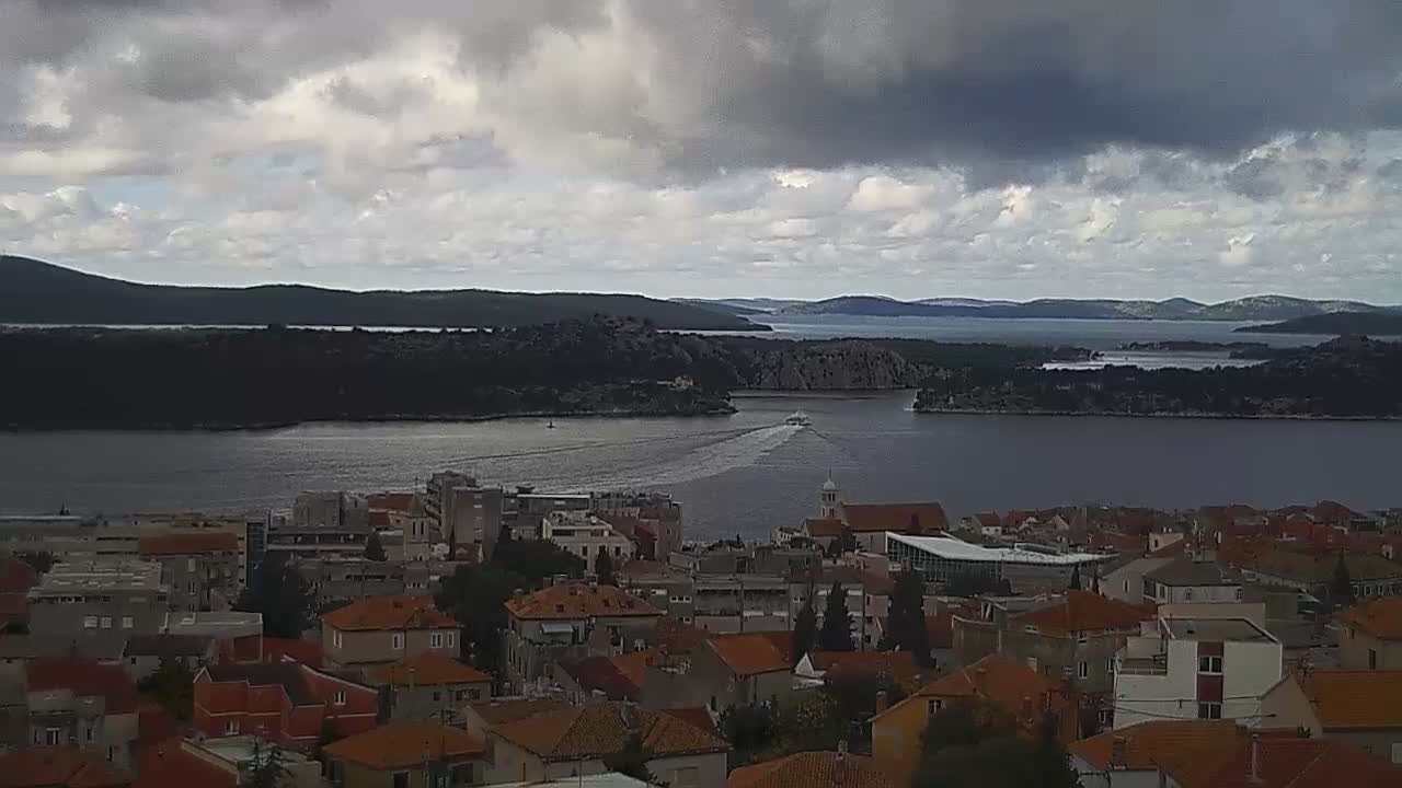 A coastal town with red-tiled roofs lines a wide body of water, through which a boat moves past distant islands under a heavily overcast sky.