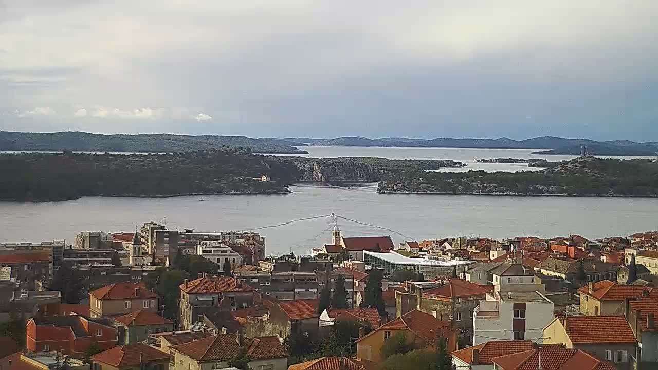 An overcast aerial view reveals a coastal town with numerous red-tiled roofs beside a wide waterway dotted with wooded islands and a small boat leaving a wake on the water.