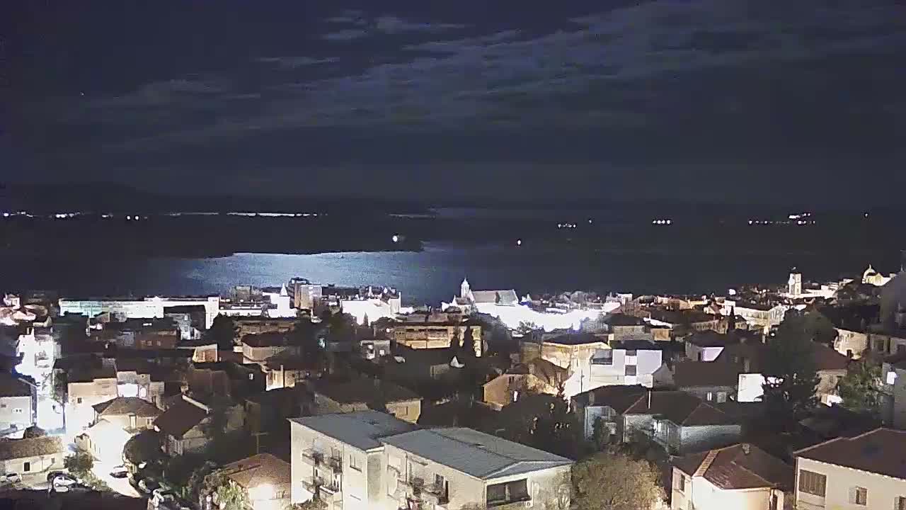An elevated view captures an illuminated town situated along a body of water, with city lights reflecting on the surface under a cloudy night sky.