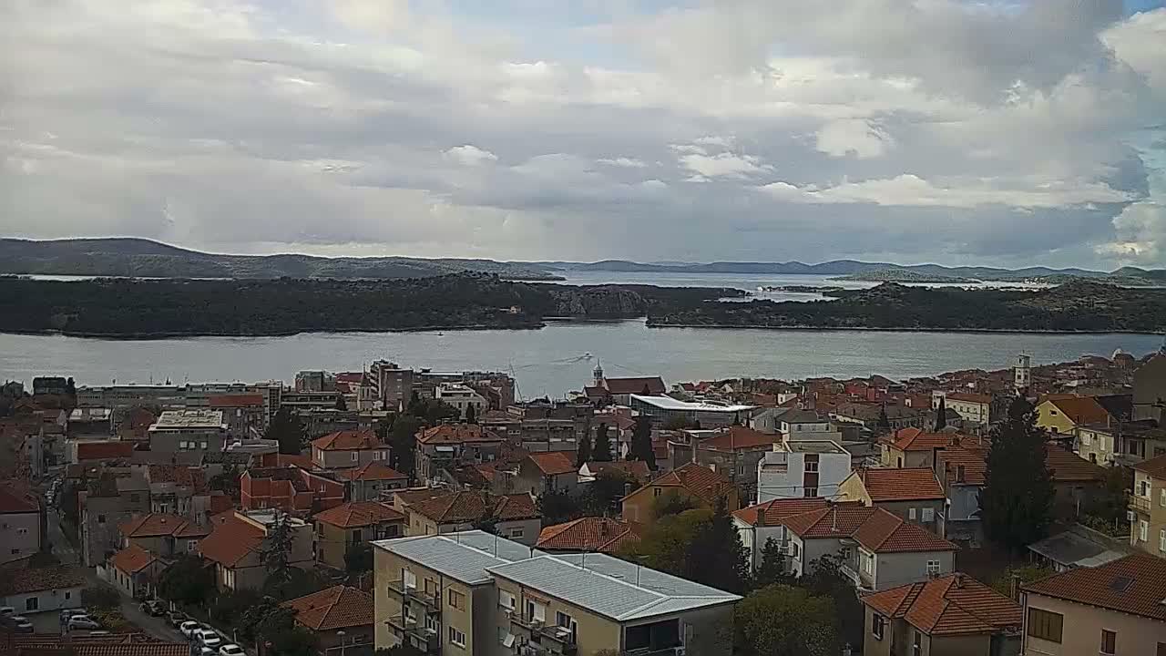 An elevated view reveals a coastal city with densely packed buildings, many featuring red-tiled roofs, bordering a wide body of water with islands and distant hills, all under a heavily overcast sky.