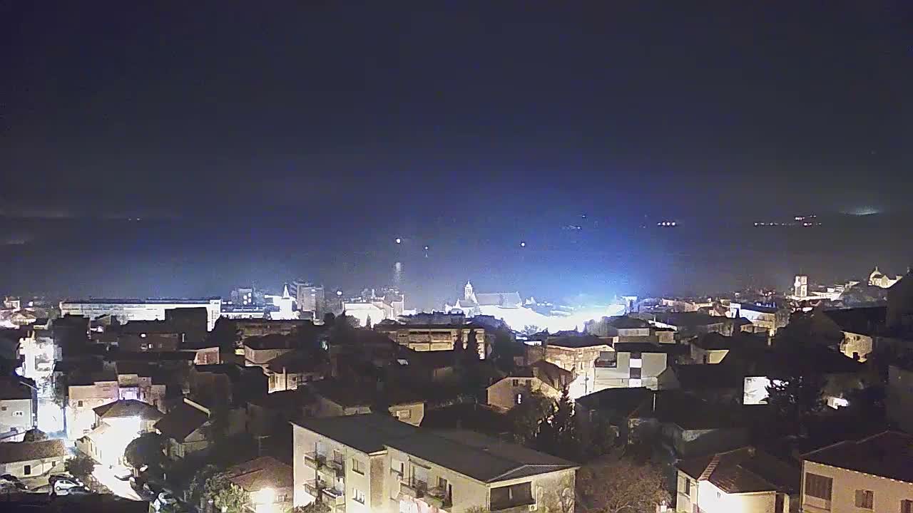 A clear night sky hangs over an urban area with numerous illuminated buildings in the foreground and midground, featuring a dramatic bright blue flash of light on the distant horizon above a body of water.