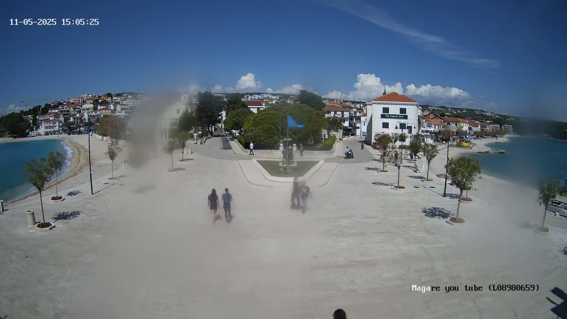 A hazy, sunny day shows a town square overlooking a calm bay, with several people walking around a central fountain and buildings lining the shore.