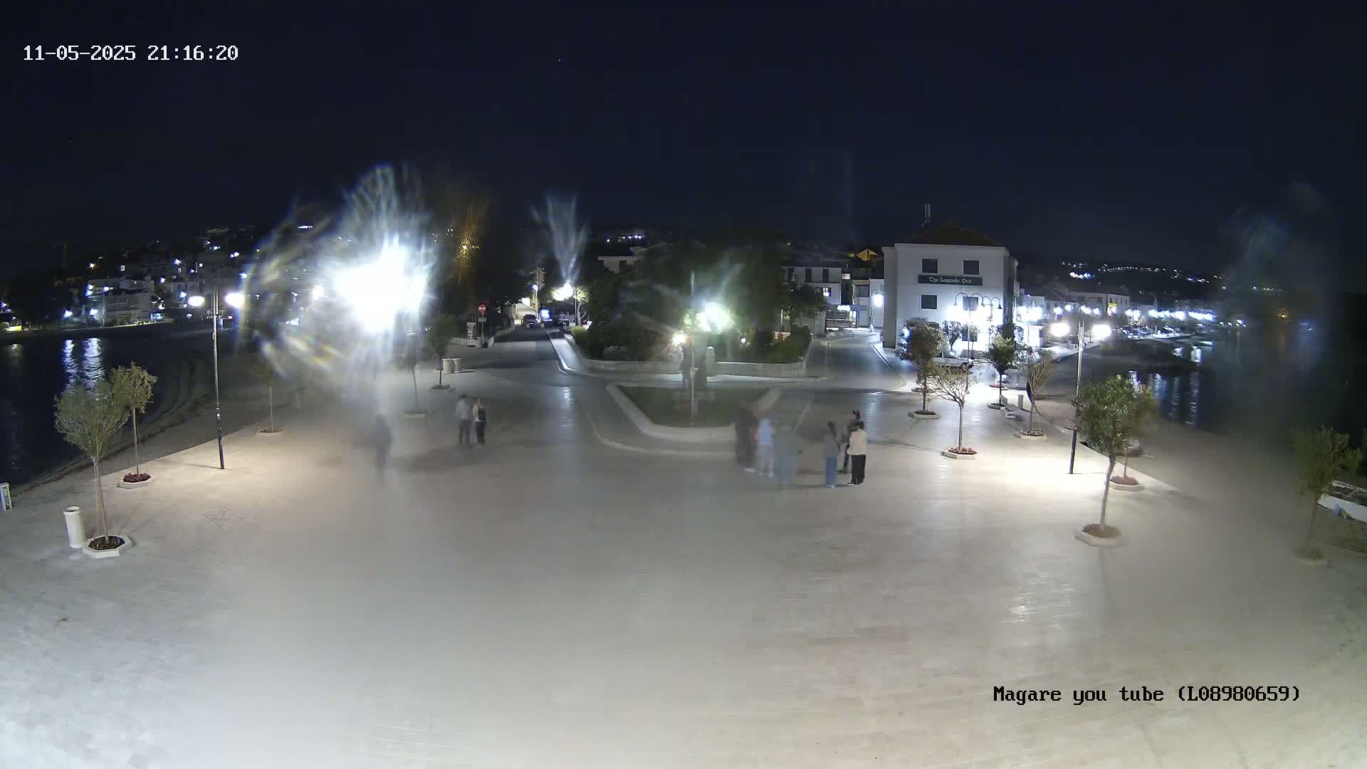 A nighttime view of a town square near a waterfront, illuminated by streetlights, with several people gathered in the square and small trees lining the paved area.
