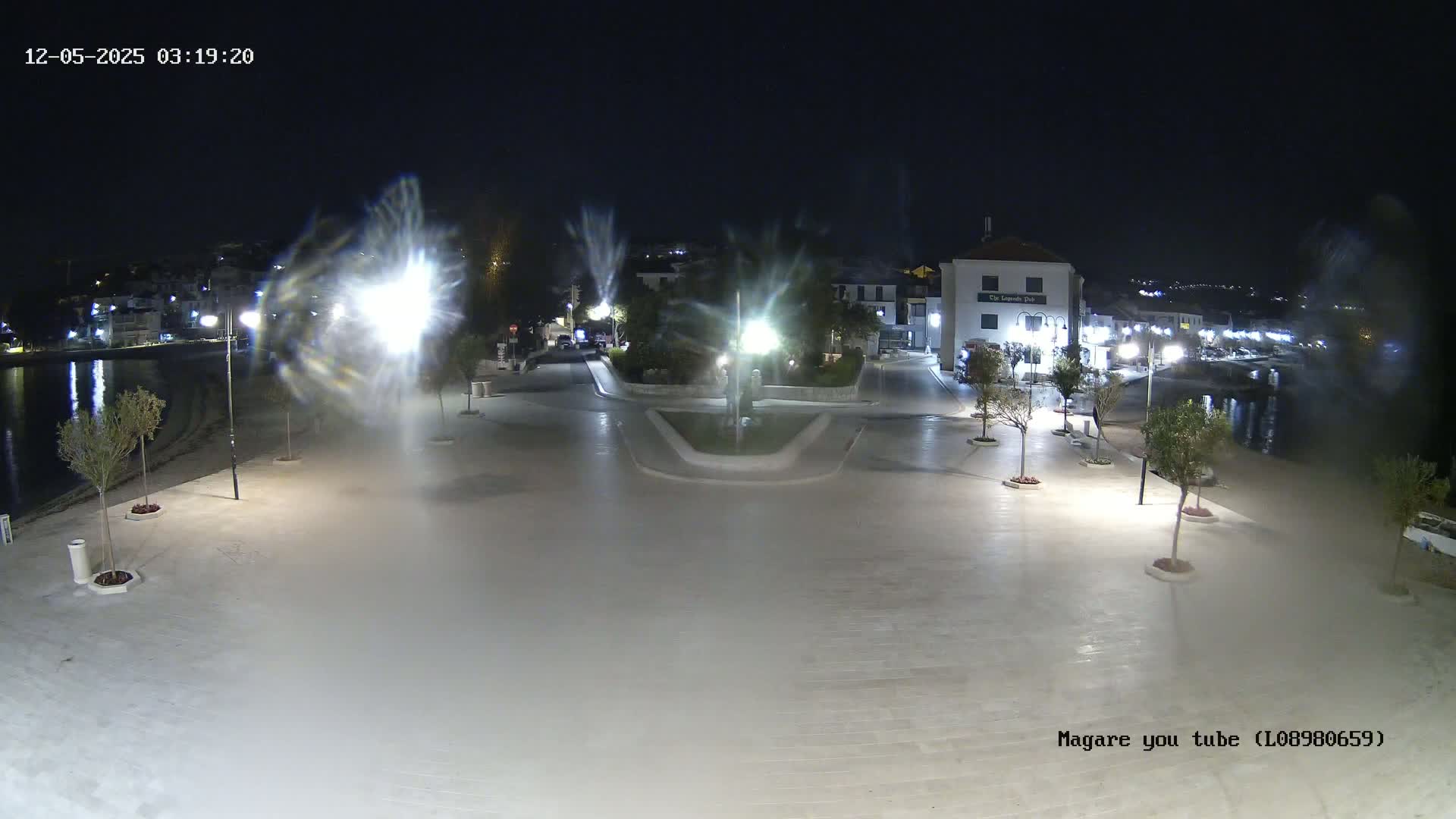 A nighttime view of a town square next to a calm body of water, illuminated by streetlights, with trees lining the paved area.