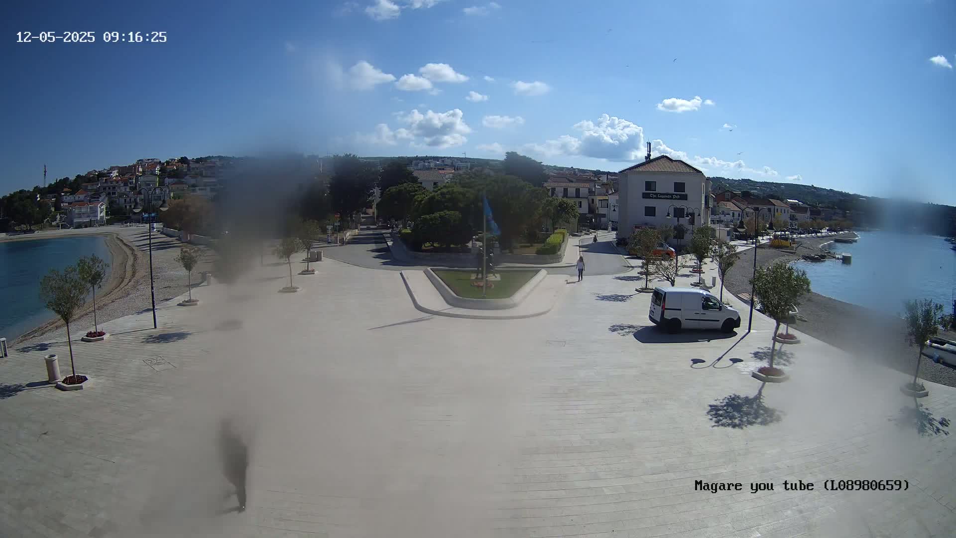 A sunny day reveals a seaside town square with a white van parked, people walking, and olive trees lining a paved area next to a calm, blue bay.