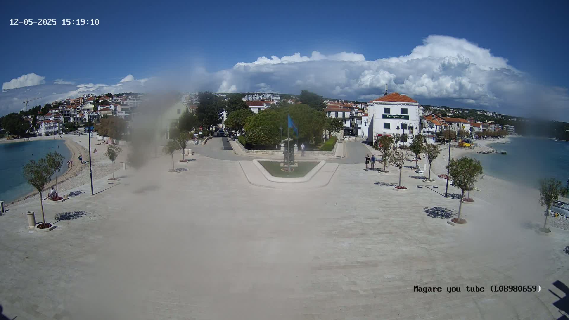 A paved town square overlooks a calm bay with a beach, under a partly cloudy sky.