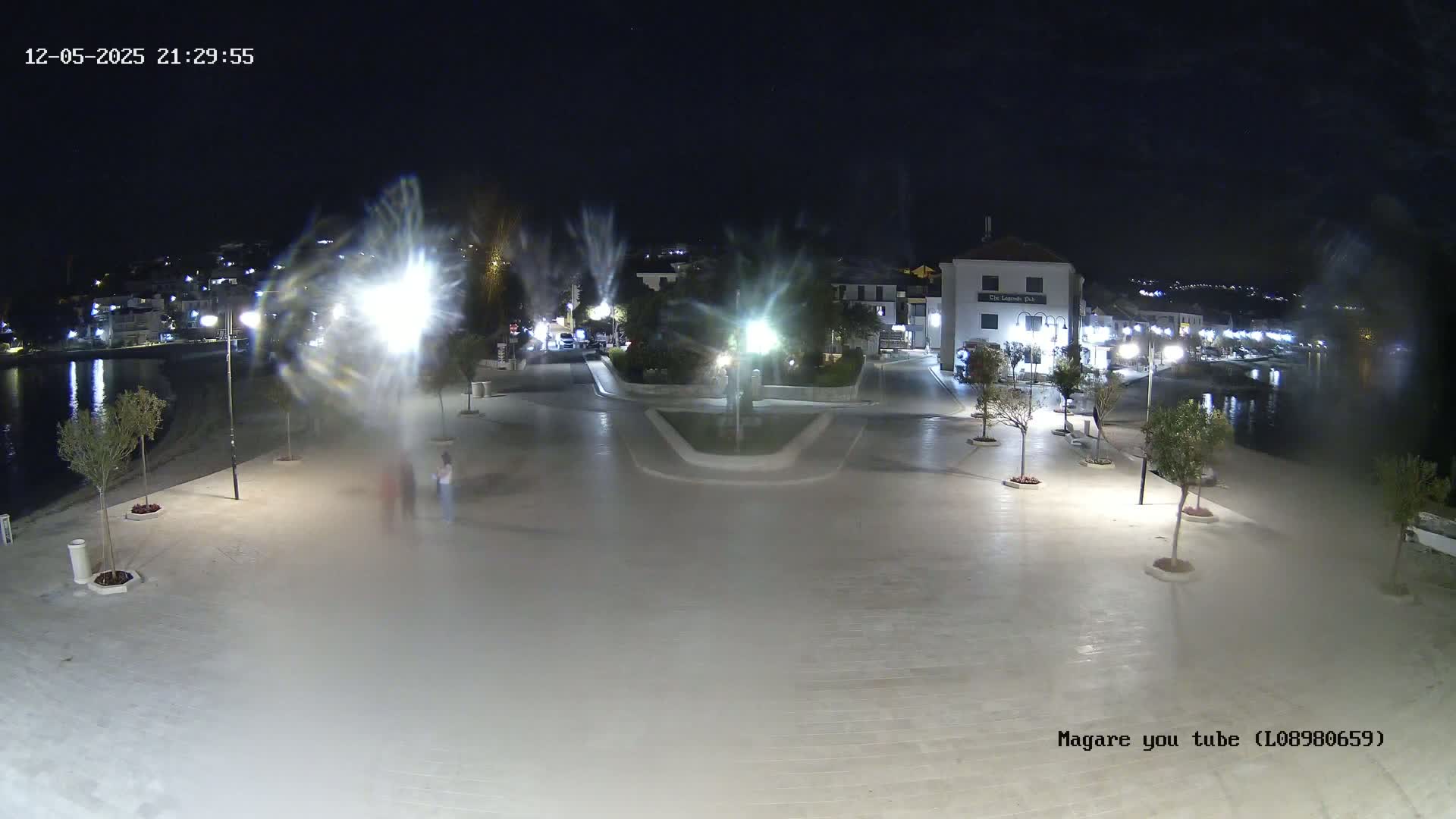 A nighttime, outdoor view of a town square near a body of water, illuminated by streetlights, with a few people walking and some trees planted around the perimeter.