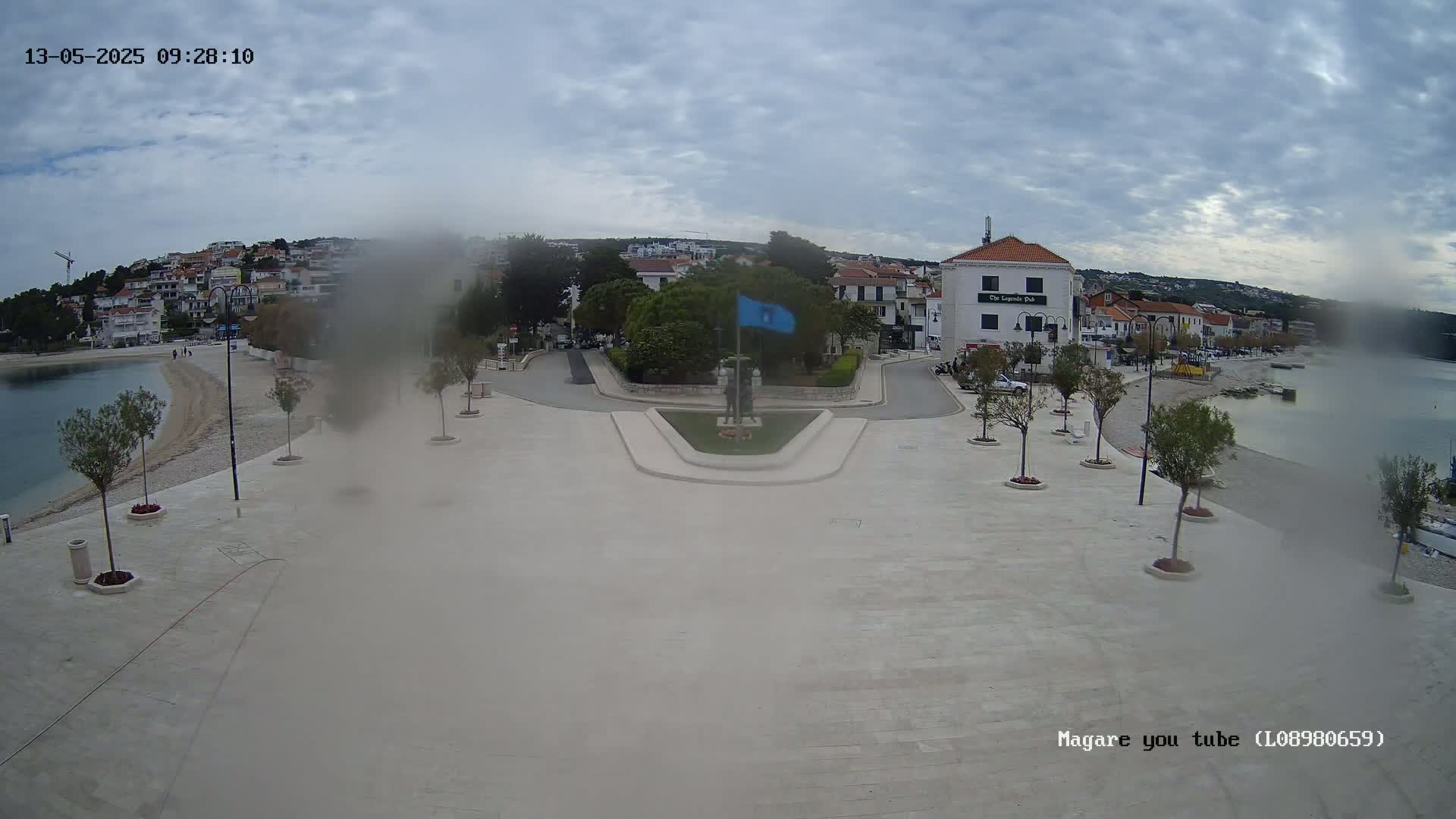 A mostly clear day reveals a town square overlooking a calm bay, with a monument at its center and small trees lining the paved area.